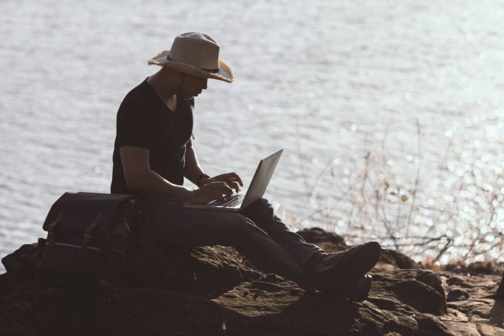 Man in straw hat using laptop on rocky lakeside, with a scenic water backdrop and bright sunlight.