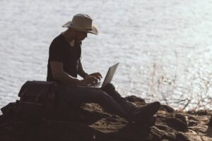 Man in straw hat using laptop on rocky lakeside, with a scenic water backdrop and bright sunlight.