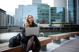 A woman using a laptop while sitting on a bench by a city waterfront, modern skyscrapers in the background.