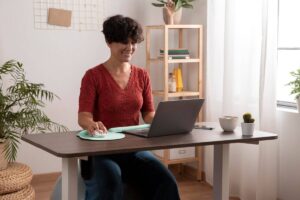 A woman in a red top smiling while working on a laptop at a home office desk.