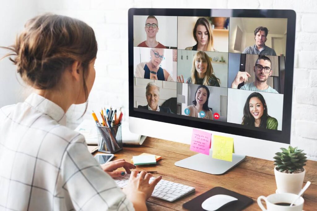 Woman in a white shirt attending a virtual meeting with multiple participants on her computer screen.