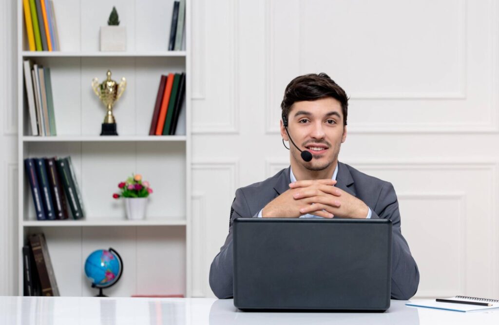 Man in a headset sitting at a desk with a laptop, in a room with a bookshelf.