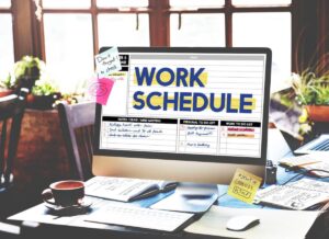 Computer screen displaying a detailed work schedule with sticky notes, in a cozy office setting with coffee and sunlight.