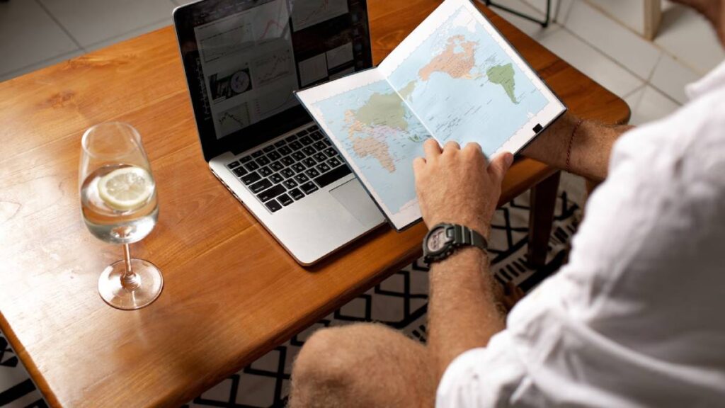 Man at a table planning a trip using a laptop and a world map, with a glass of water beside him.