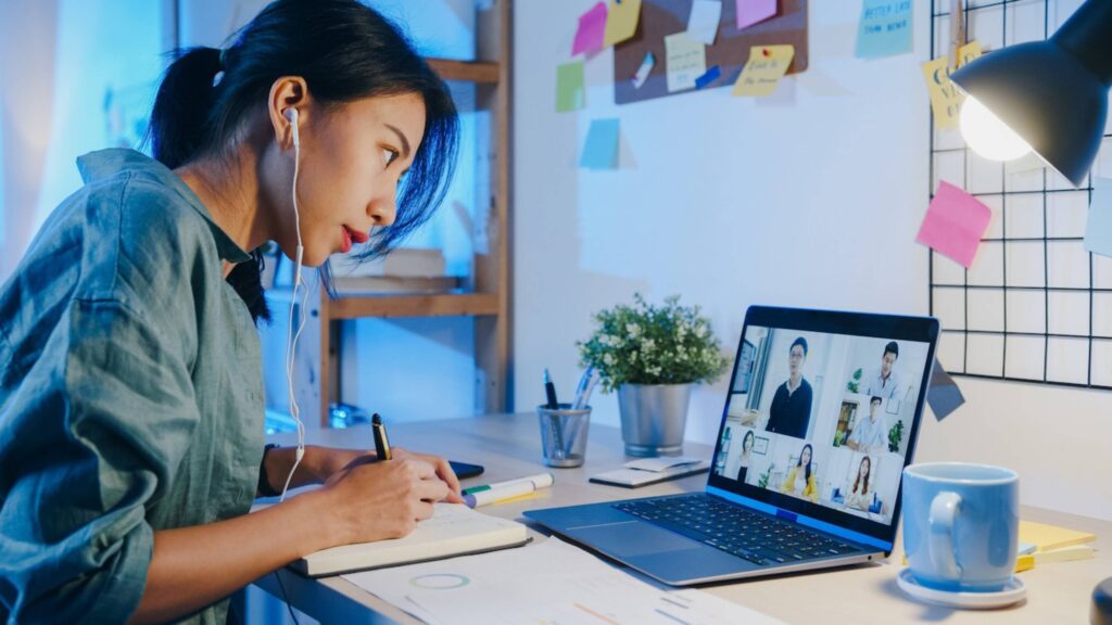 A woman participating in a virtual meeting on her laptop while taking notes at her desk filled with stationery and a cup.