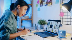 A woman participating in a virtual meeting on her laptop while taking notes at her desk filled with stationery and a cup.