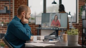 Man adjusting earpiece during a video call with a woman on the monitor in a modern office setting.