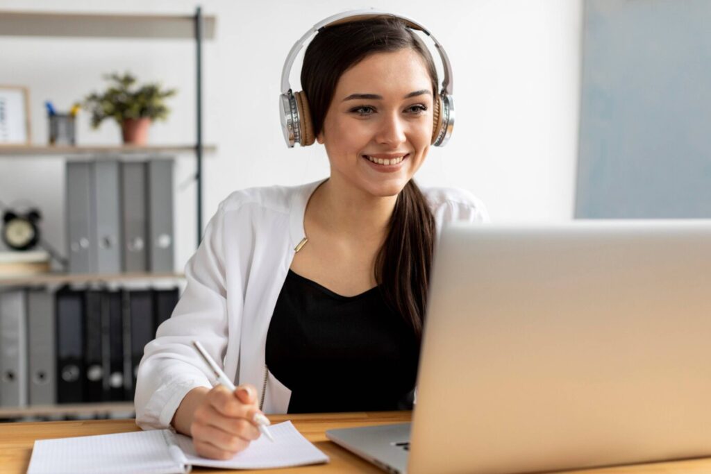 Young woman wearing headphones, smiling while using a laptop in an office setting.