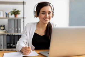 Young woman wearing headphones, smiling while using a laptop in an office setting.