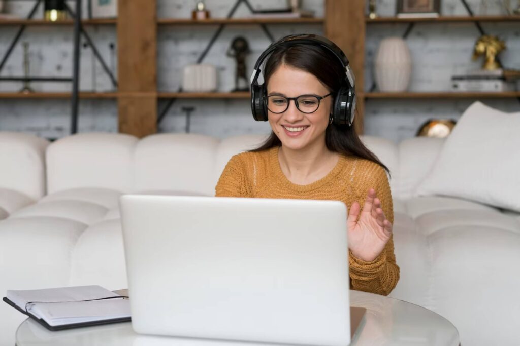 A smiling woman with headphones using a laptop, gesturing with her hand, in a cozy living room.
