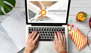 Hands typing on a laptop showing a certification screen, surrounded by snacks and notebooks on a wooden desk.