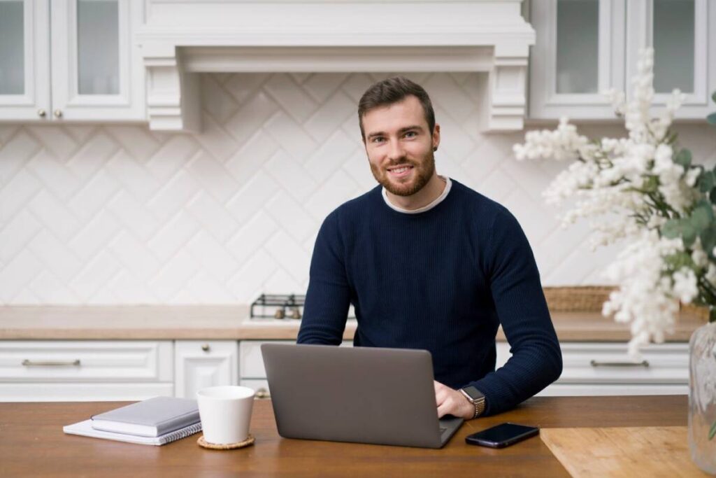 A man with a beard smiling at the camera while working on a laptop in a kitchen.