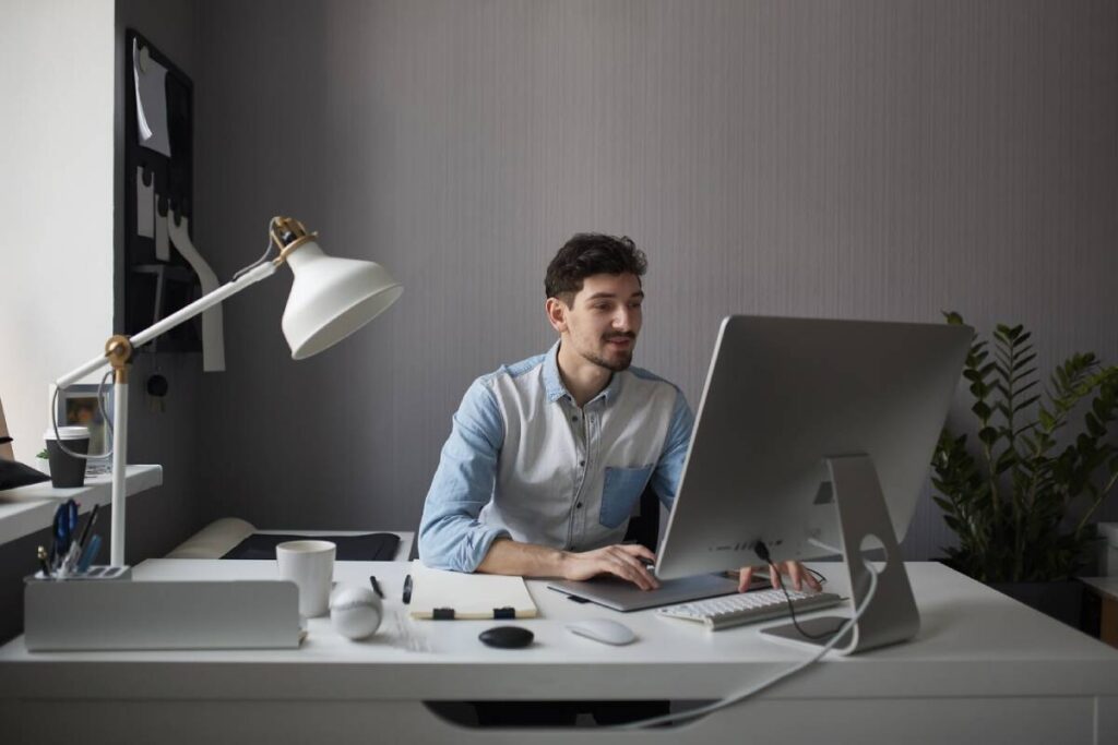Man sitting at a desk working on a computer with a desk lamp and minimalistic decor.
