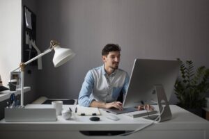 Man sitting at a desk working on a computer with a desk lamp and minimalistic decor.