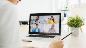 Person attending a video call with five diverse participants on a laptop screen in a bright office.