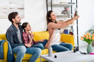 Family of three taking a selfie on a yellow sofa in a bright living room.