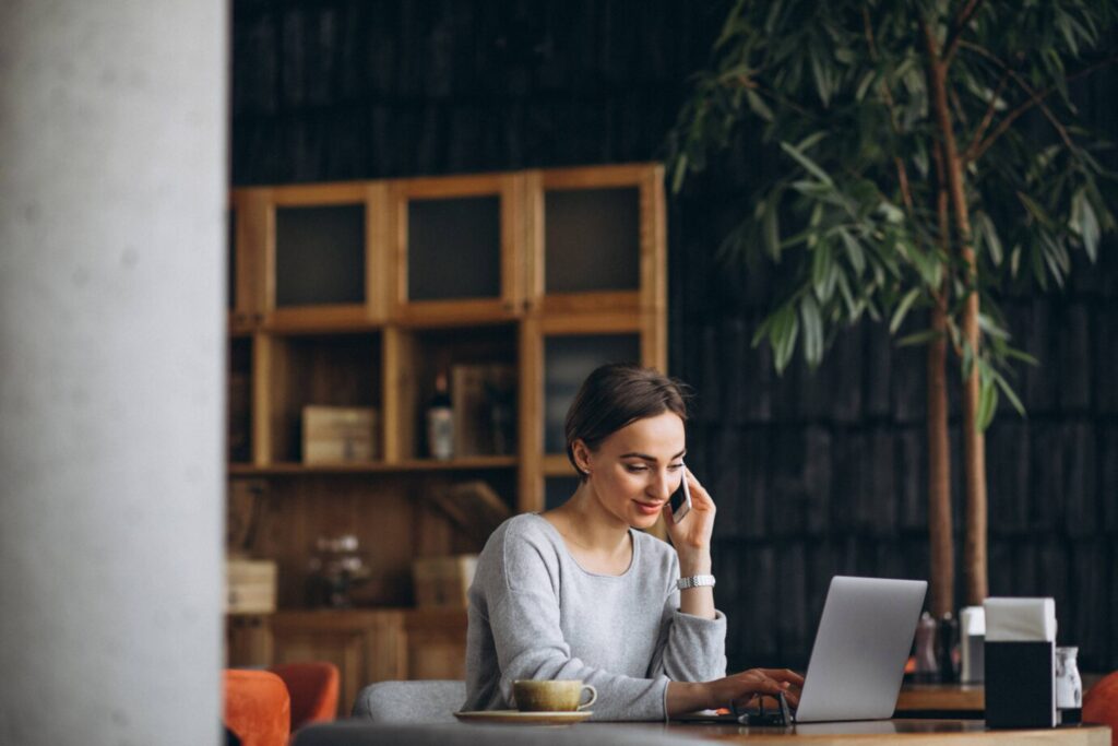 A woman smiling while using a laptop in a cozy, plant-decorated café setting.
