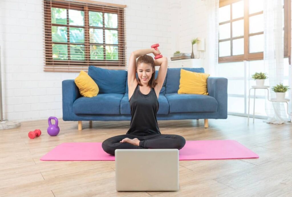 Woman exercising with a dumbbell in her living room, following an online workout on her laptop.