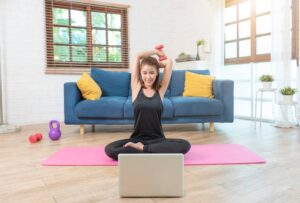 Woman exercising with a dumbbell in her living room, following an online workout on her laptop.