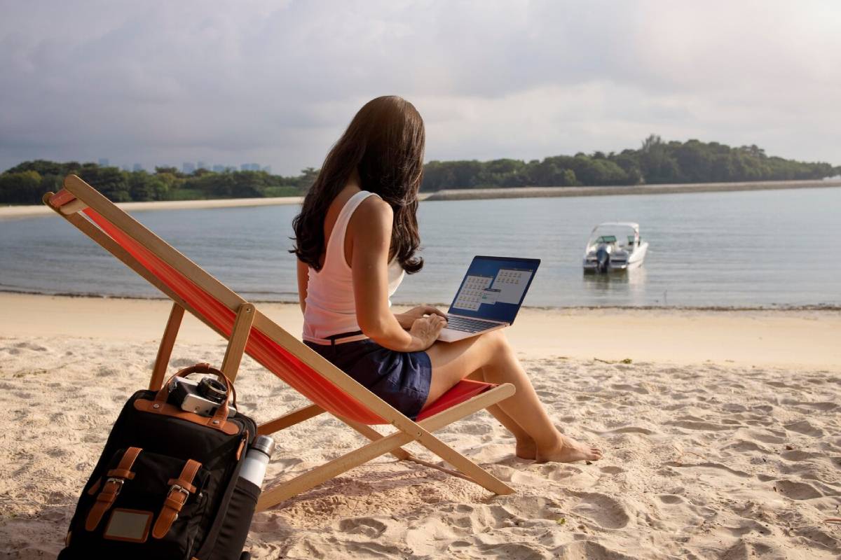Woman using a laptop on a beach chair by the sea with a boat in the background. 