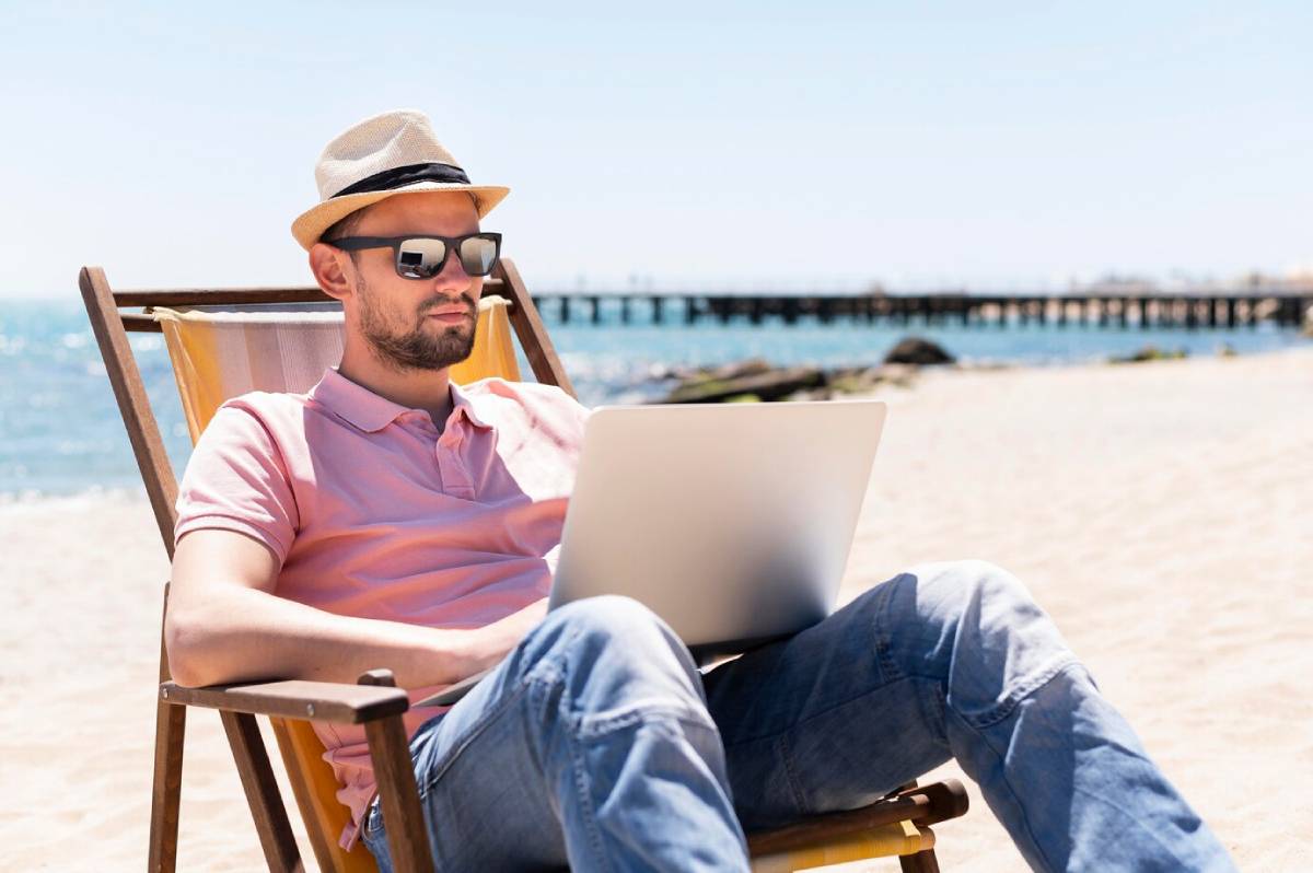 Man in sunglasses and hat working on a laptop while sitting in a deck chair on a sandy beach. 