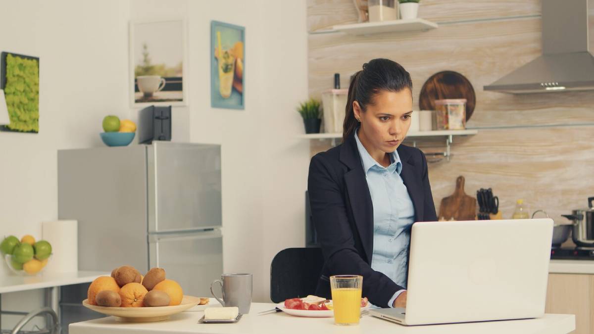 A woman in a business suit working on a laptop in a kitchen with breakfast on the table. 