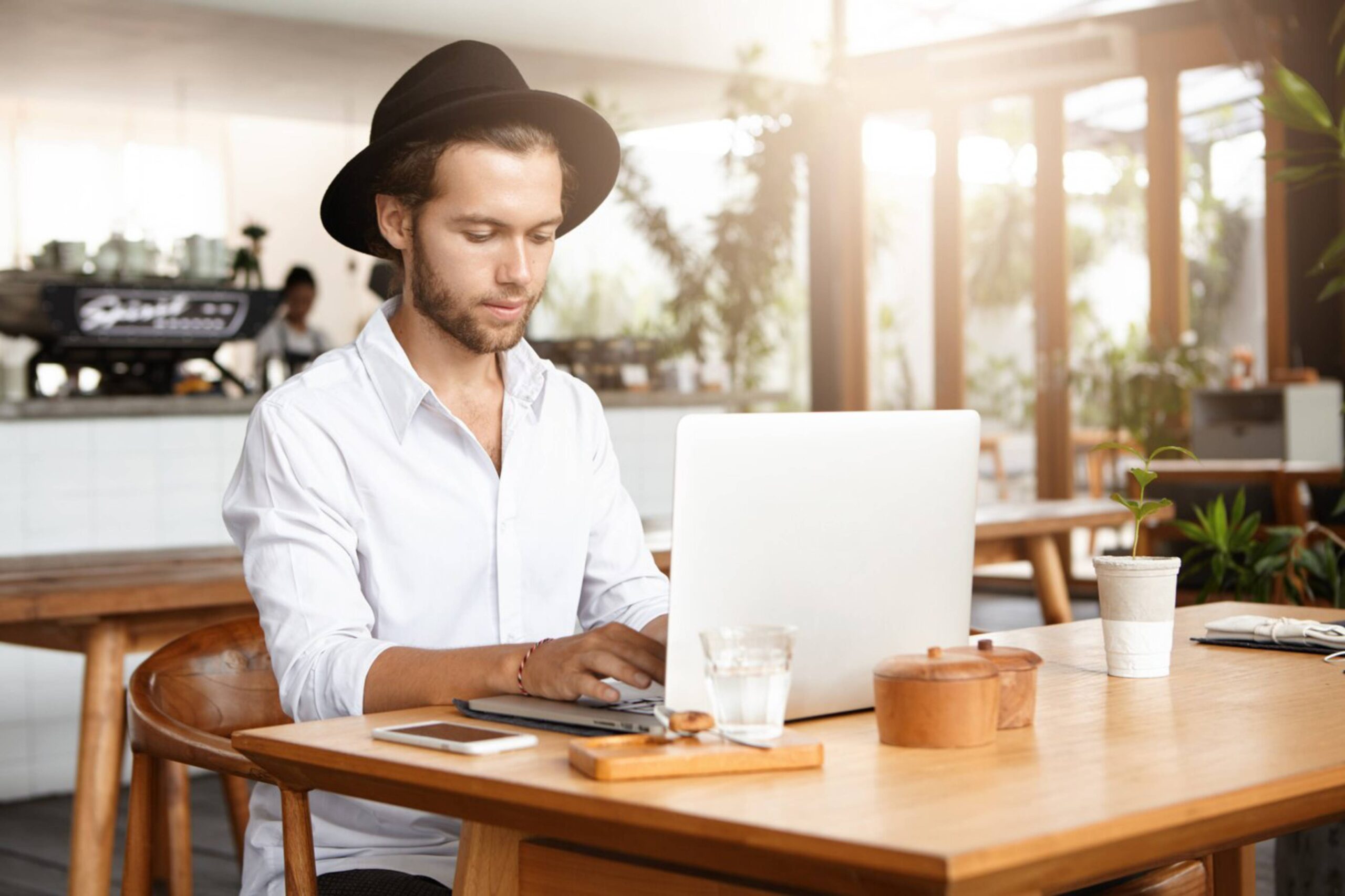 A young man in a hat typing on a laptop at a cafe table. 