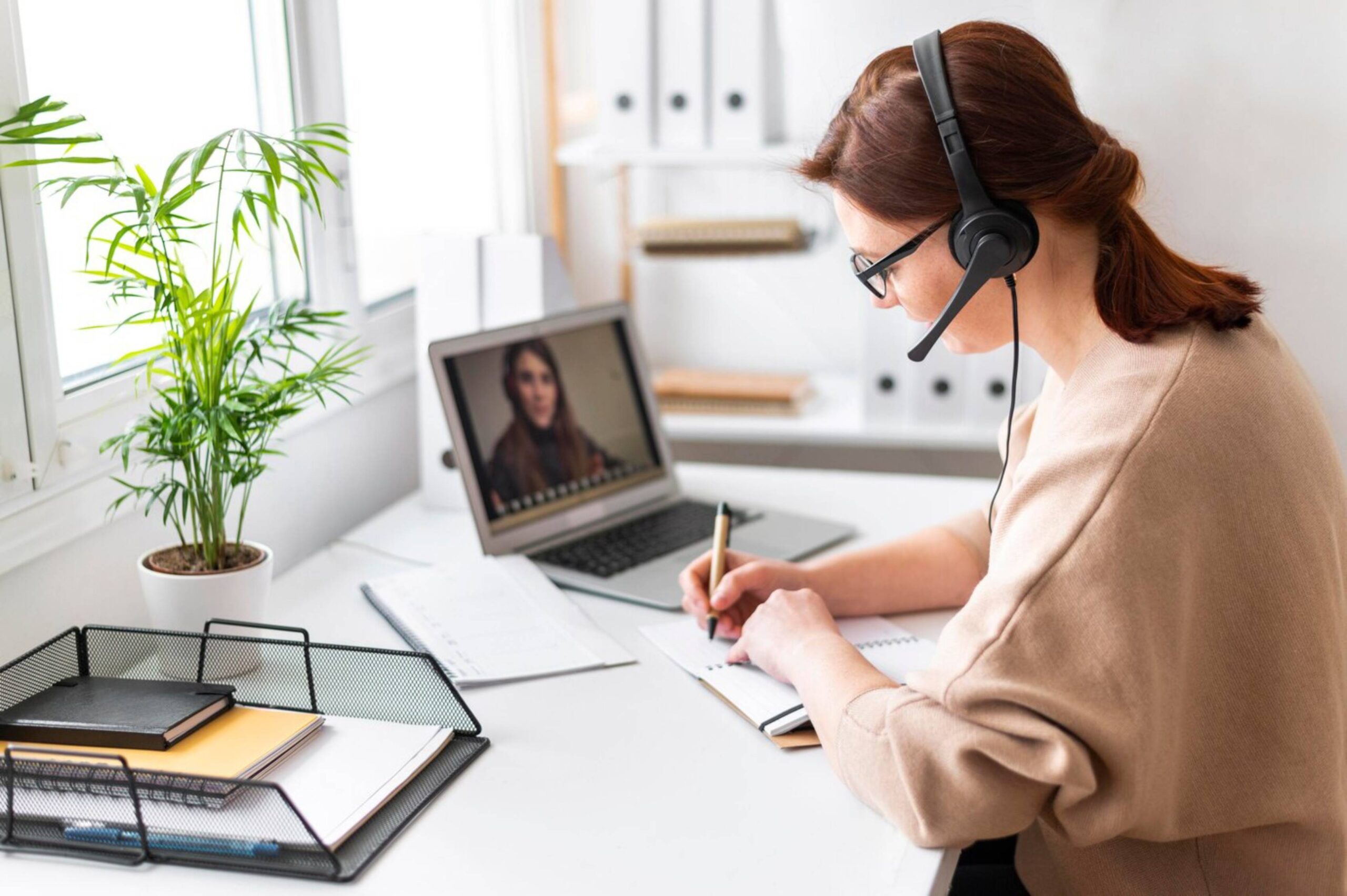  A woman with a headset participates in a video call while writing notes at her office desk.