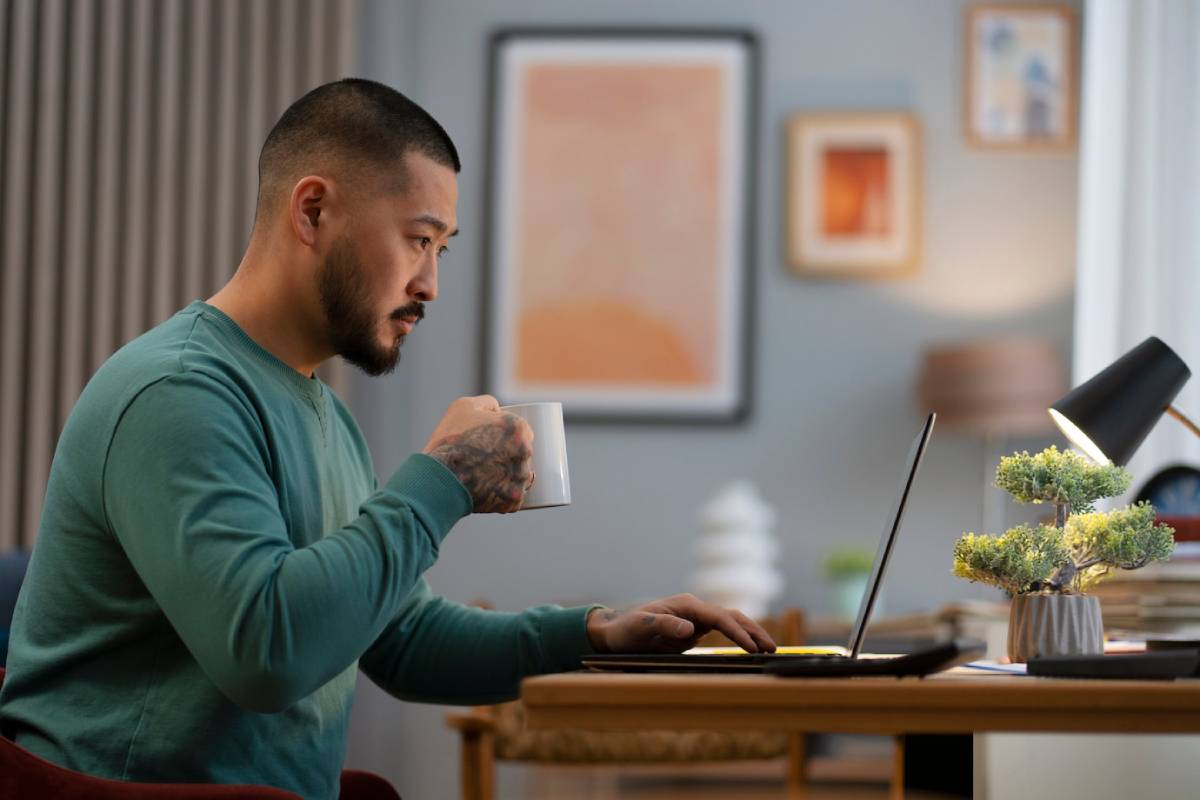 Man with tattoos working on a laptop and holding a mug in a stylish home office. 