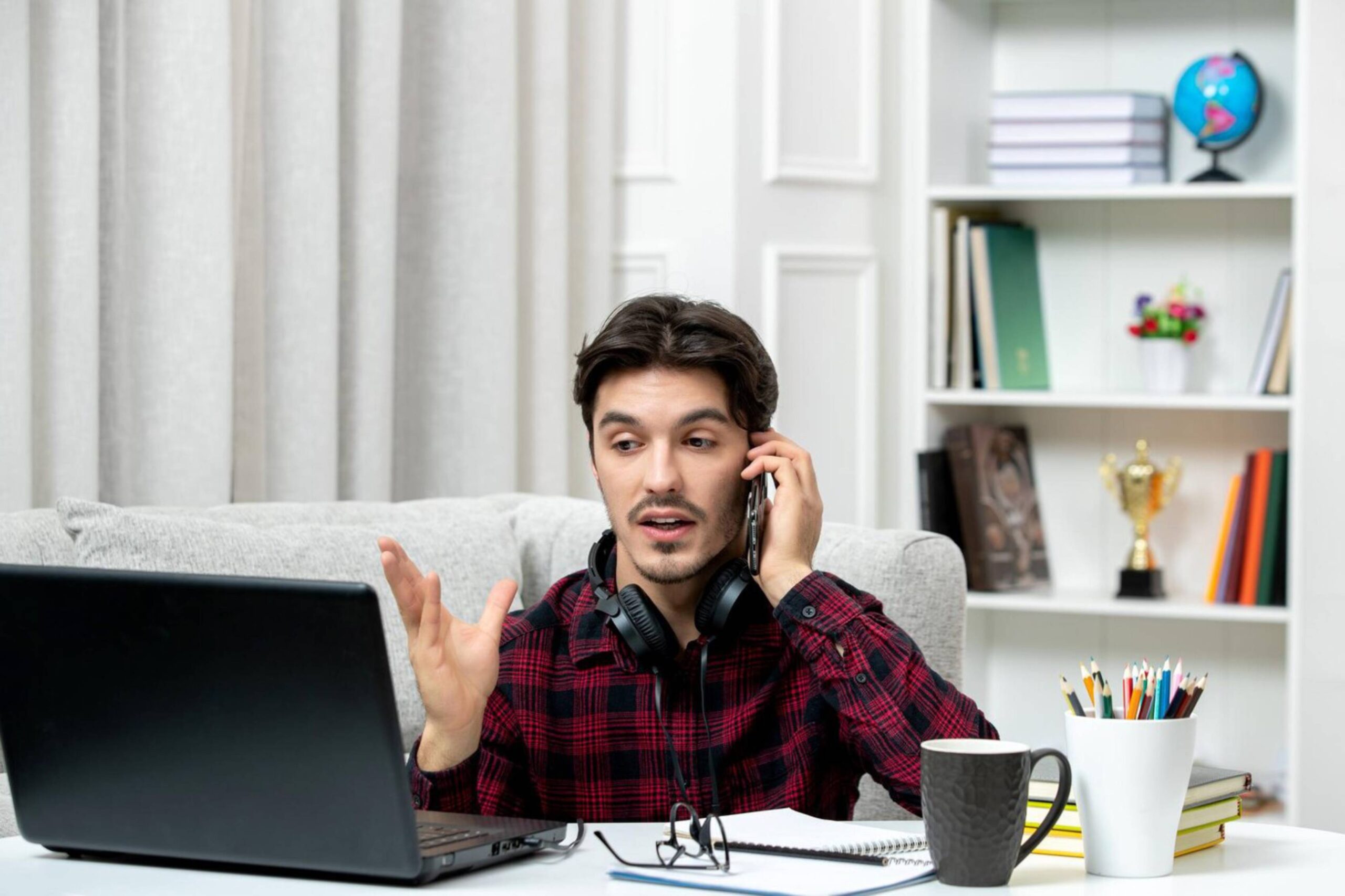 Man in plaid shirt talking on phone while working at laptop in a home office setting. 