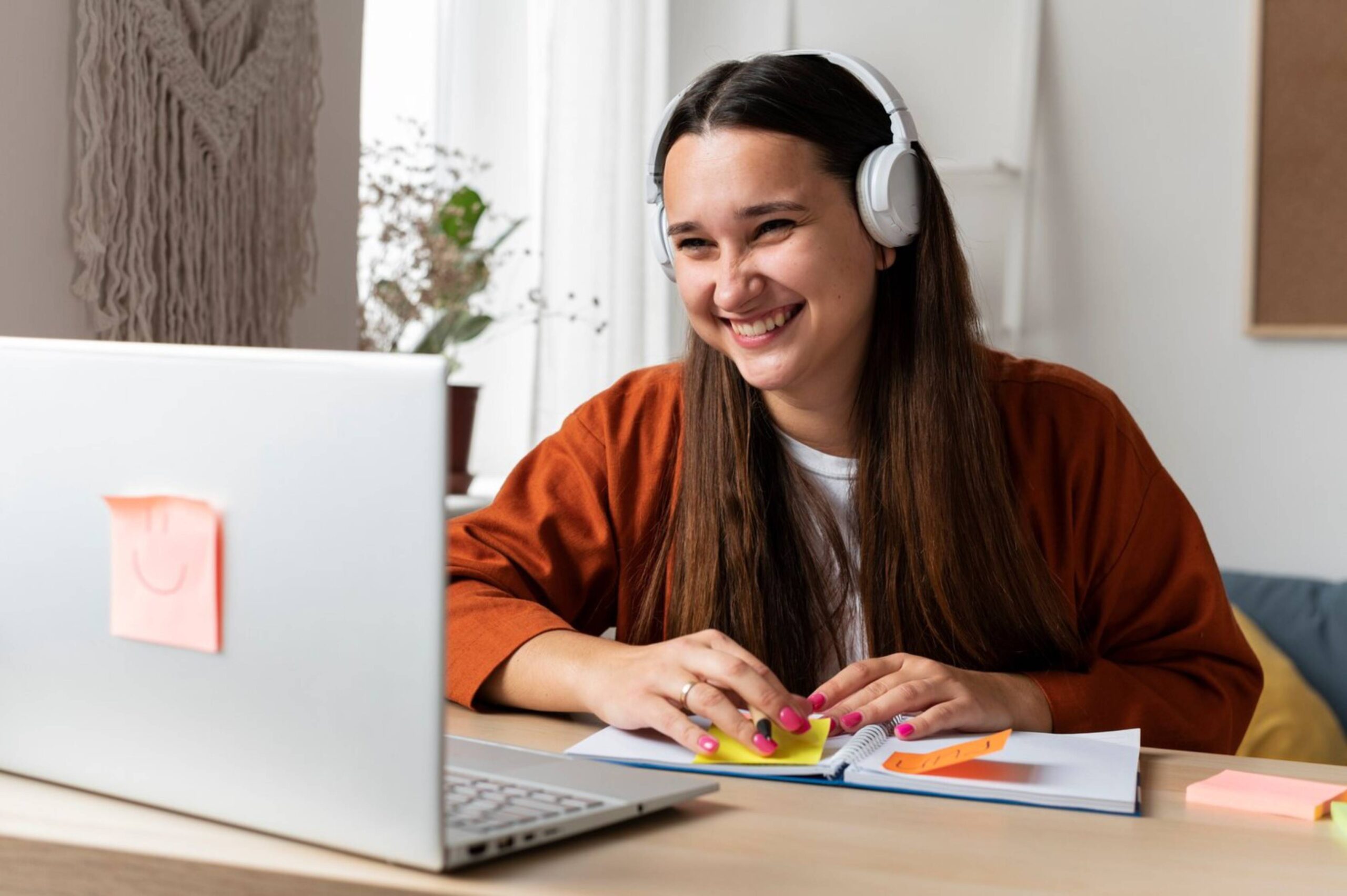  Young woman studying at home, smiling while using laptop and wearing headphones.