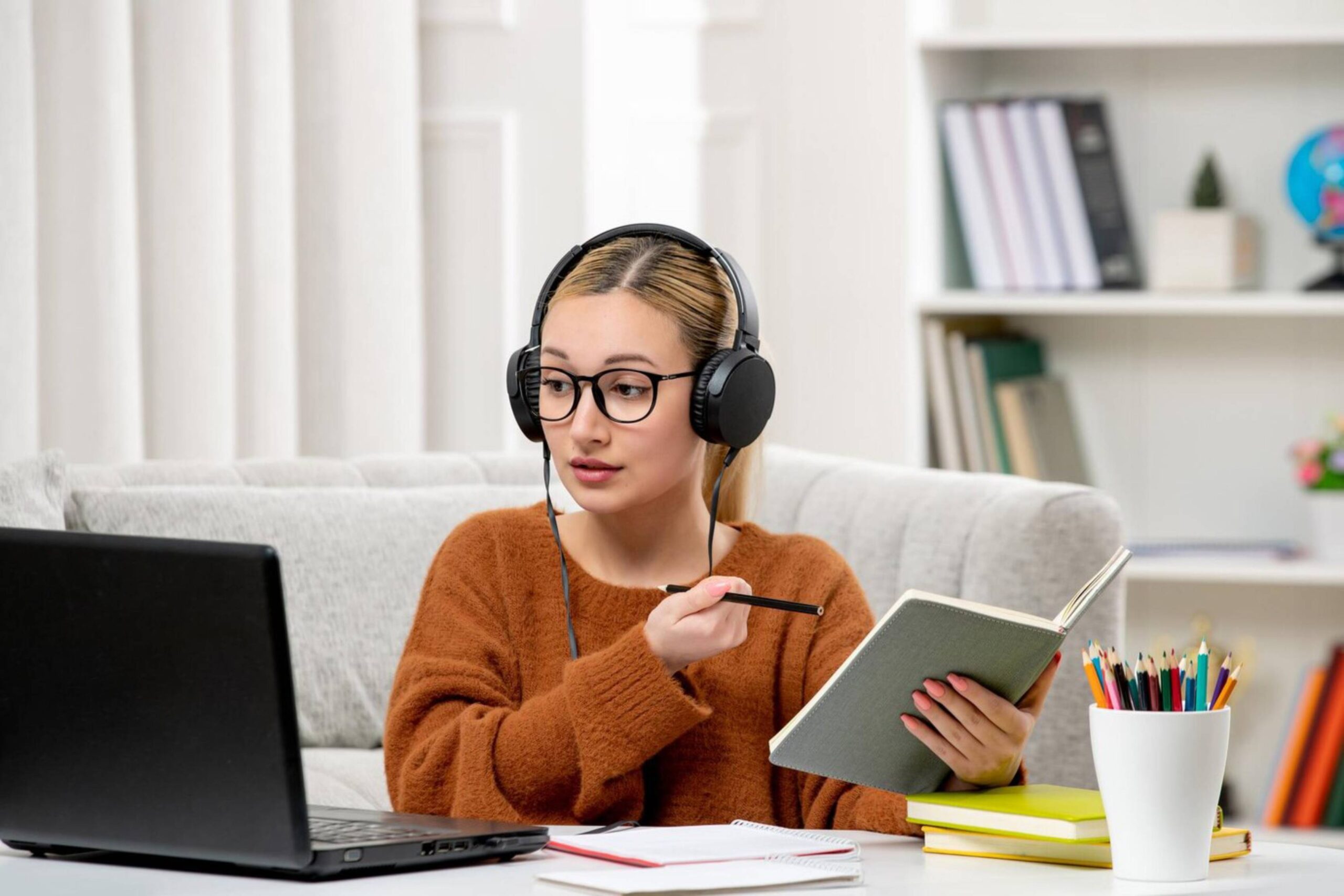 Young woman studying online with headphones and a laptop, holding a notebook and pencil in a home setting. 