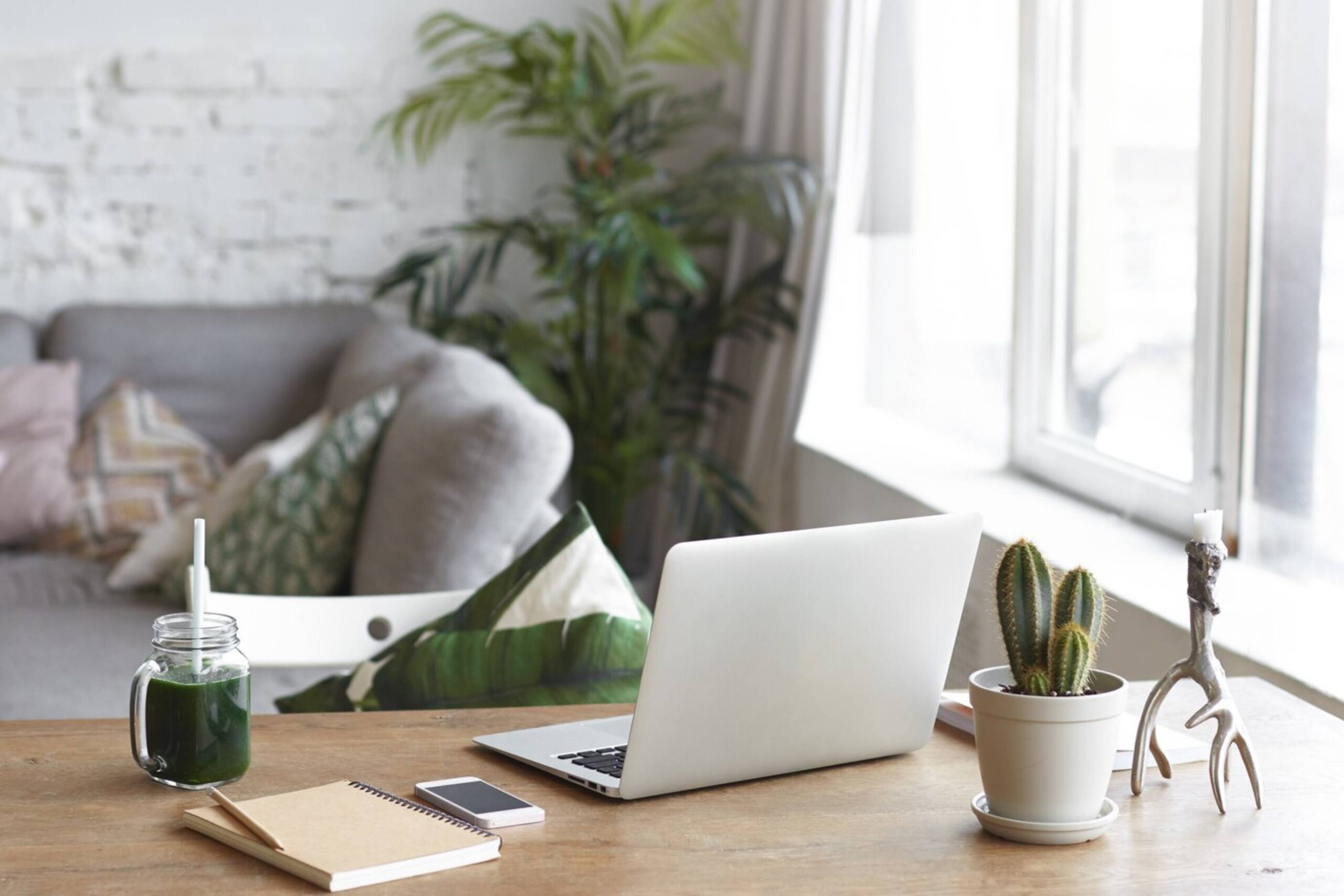  A modern workspace featuring a laptop, green smoothie, and a cactus on a wooden desk in a bright room.
