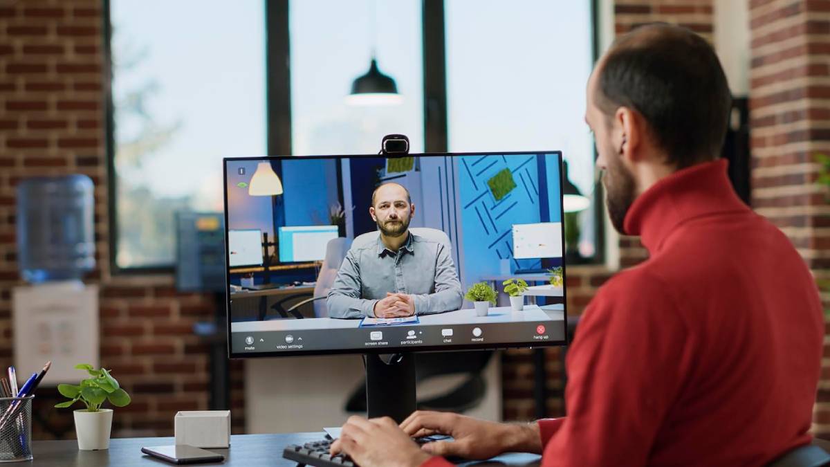  A man in a red sweater participates in a video call with another man on a large monitor in a modern office.