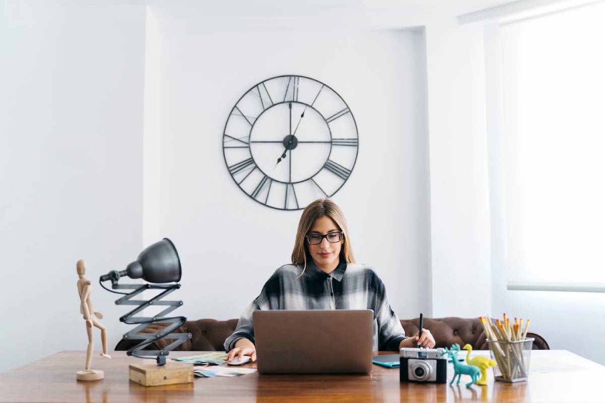 Woman working on a laptop at a tidy desk with a large clock on the wall behind her. 