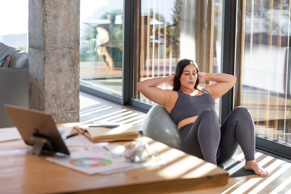  Woman exercising on a fitness ball at home, with laptop and books nearby.