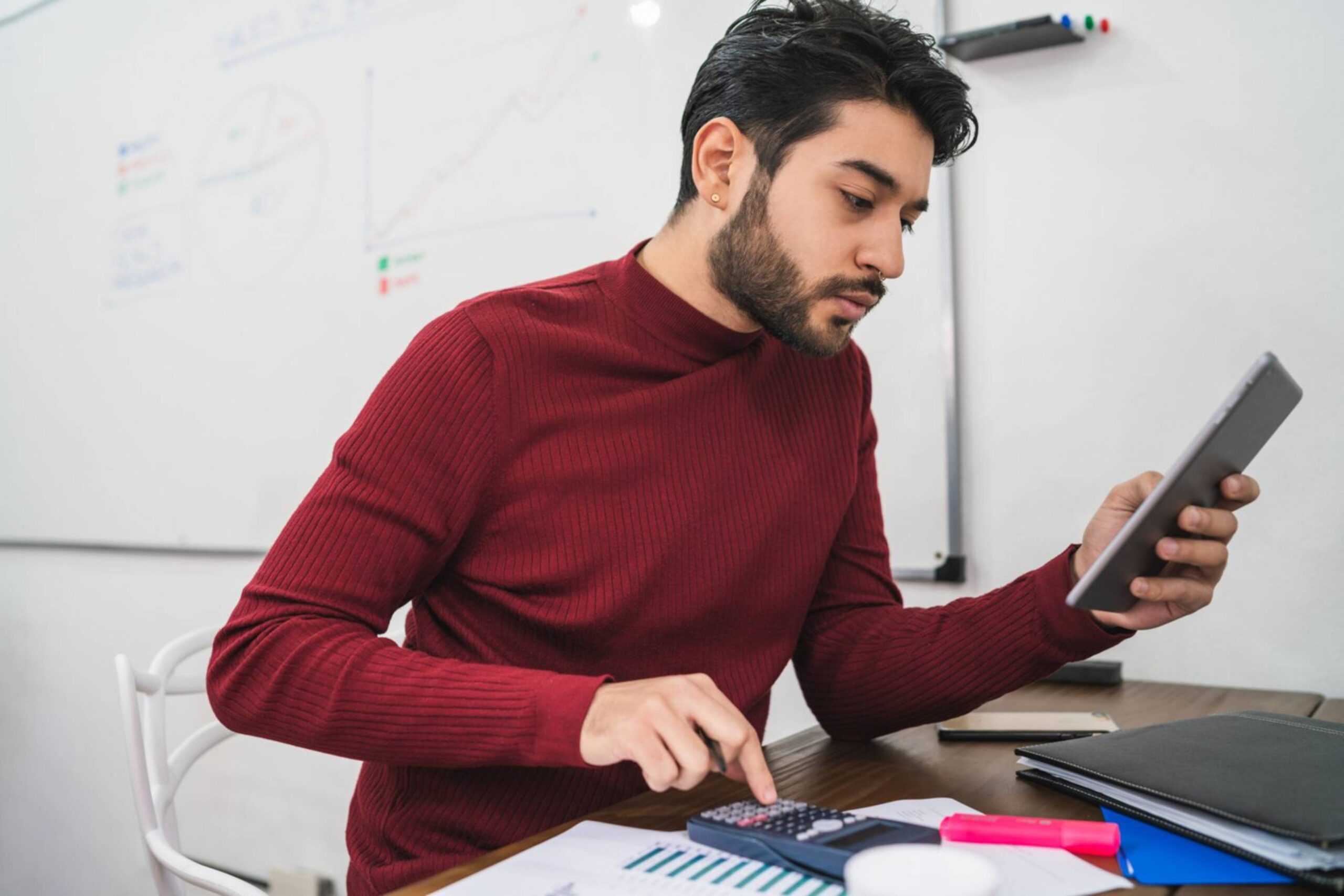  Man in red sweater analyzing data on tablet and using calculator at office desk.