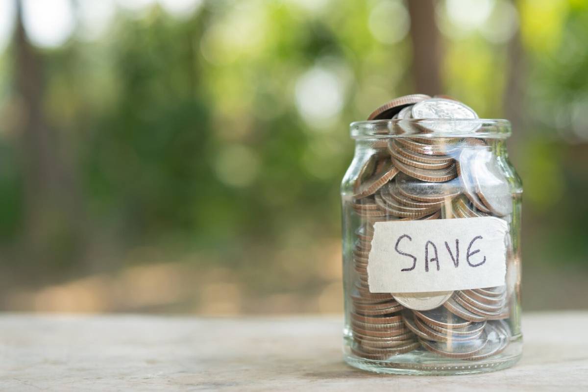 A glass jar filled with coins, labelled 'SAVE', on a wooden table. Background is a soft green blur. 