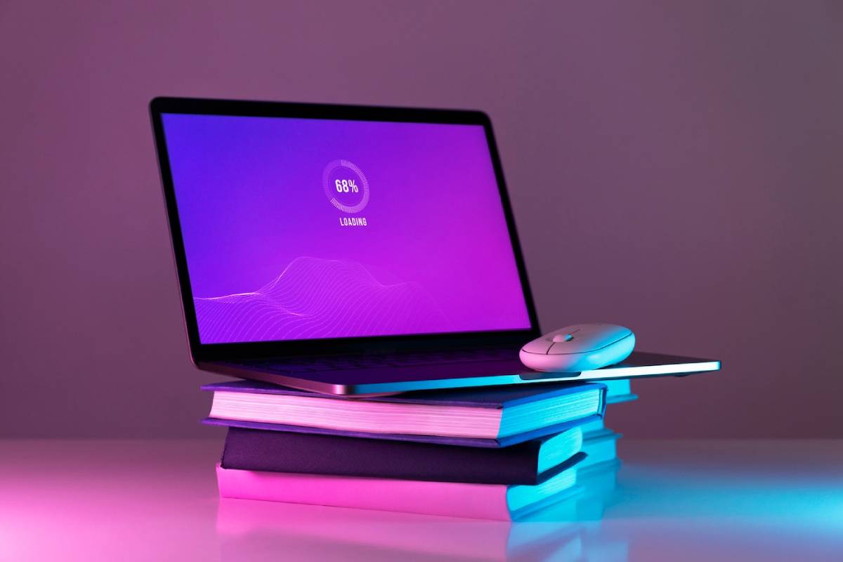  Laptop on stacked books with a loading screen and a mouse, illuminated by purple and blue lights.