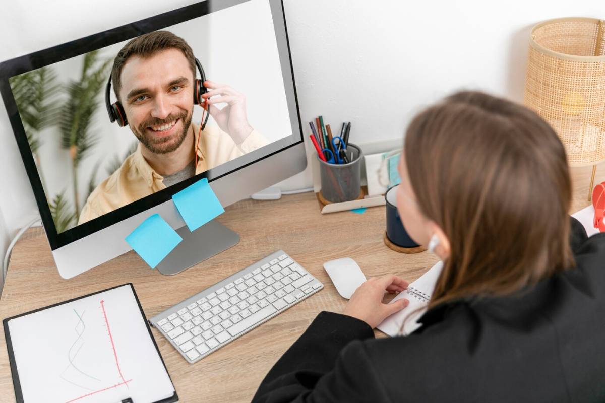  A woman at a desk talks to a smiling man on a computer screen; office setting with stationery and decorative plant visible.