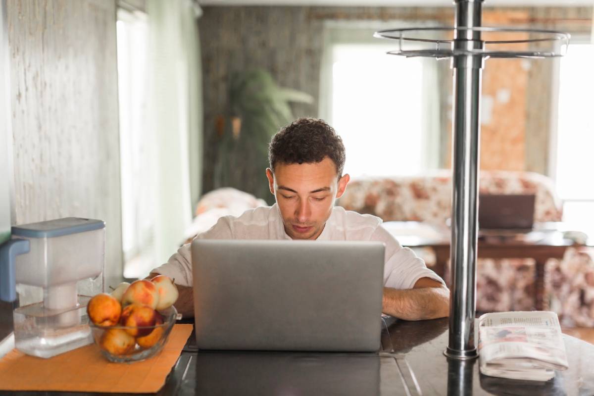  Man working on a laptop in a kitchen with a bowl of fruit nearby.