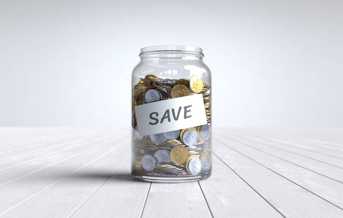  Clear glass jar filled with various coins, labelled with a save tag, placed on a wooden surface.