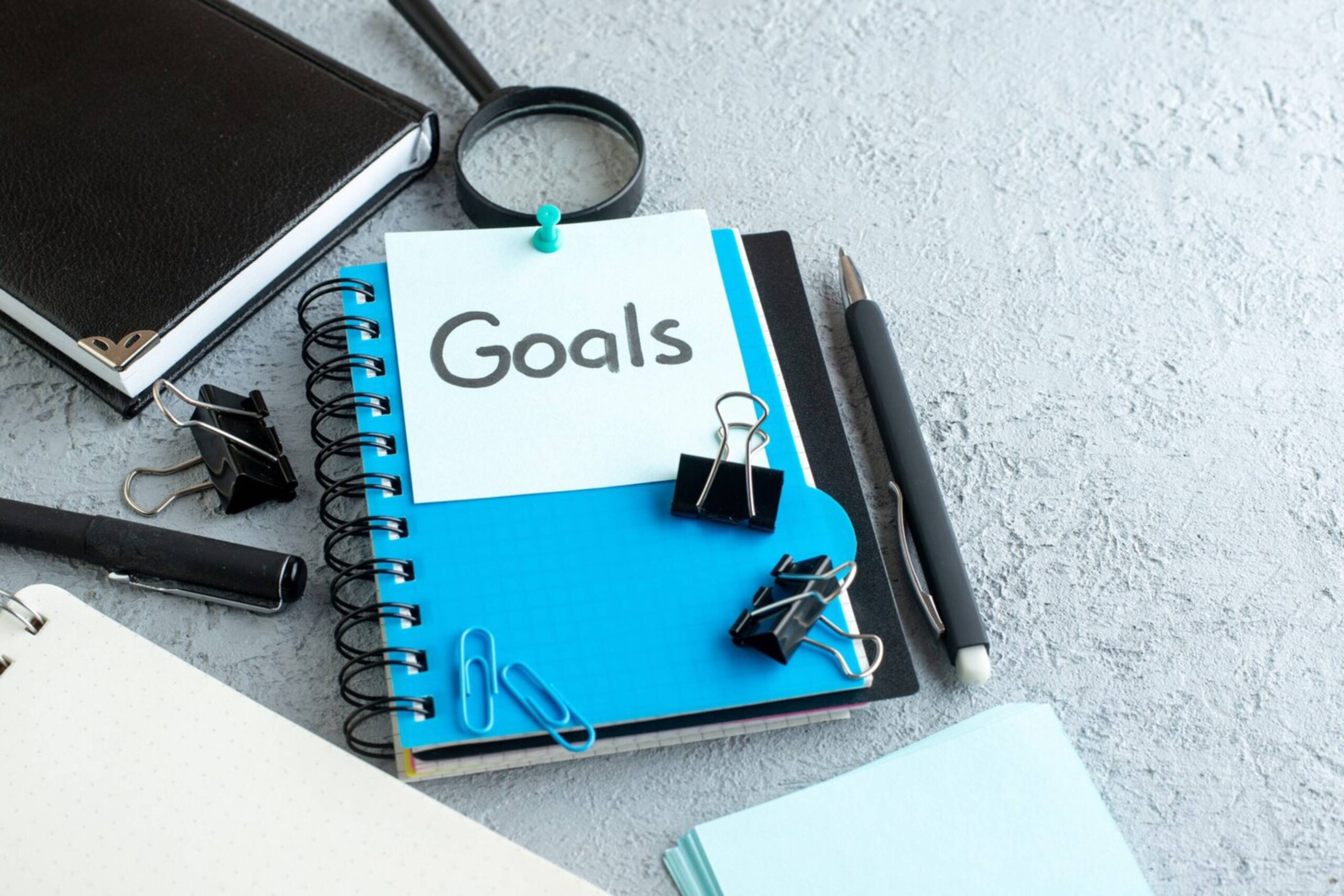  Notebook labelled 'Goals' with pens, a magnifying glass, and stationery on a textured grey surface.