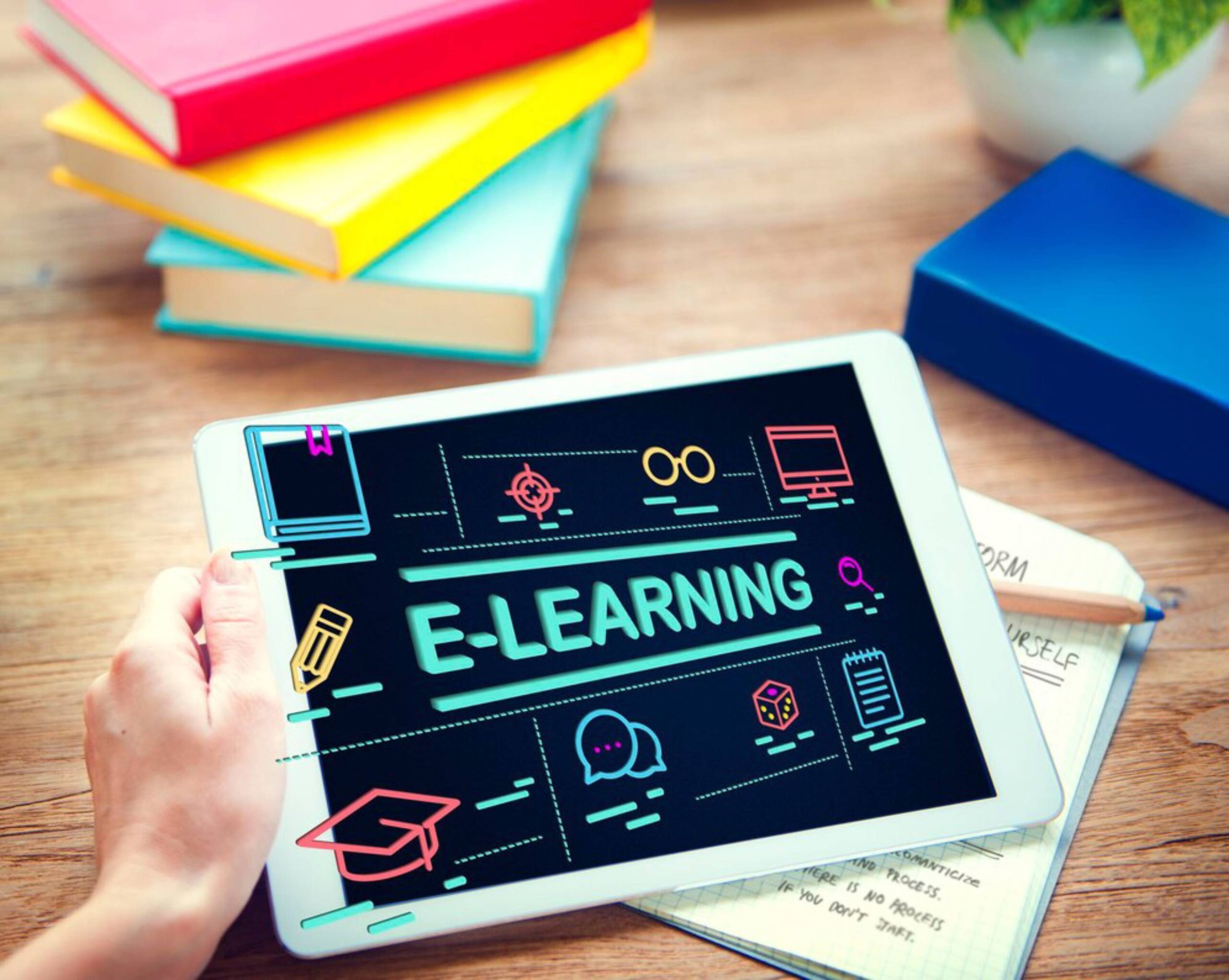 A person's hand holding a tablet displaying colorful e-learning graphics, surrounded by books and stationery on a wooden desk. 