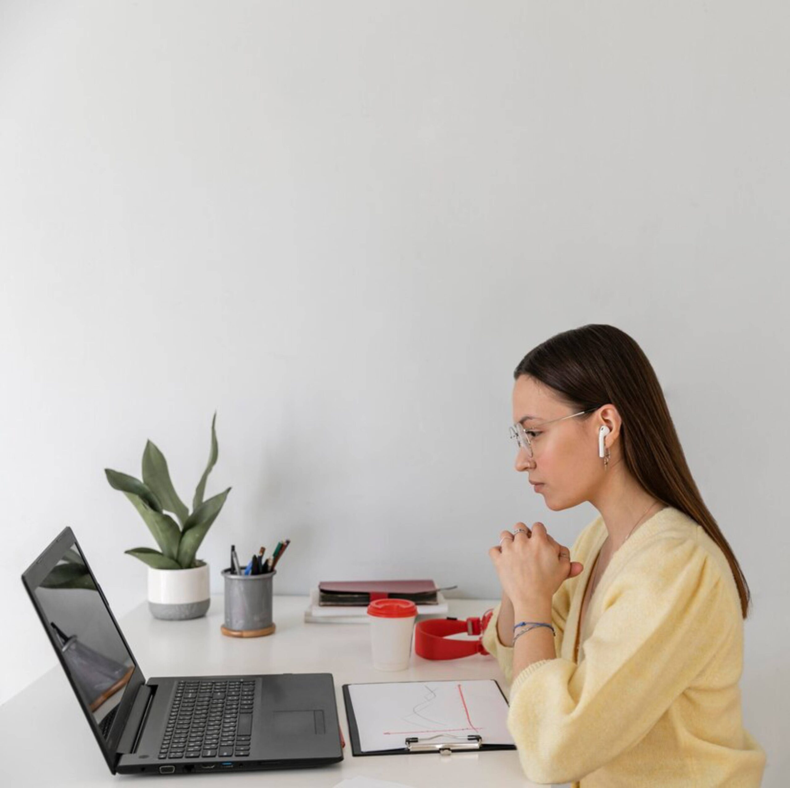  Woman in glasses working intently at a laptop, with plants and office items on a white desk.