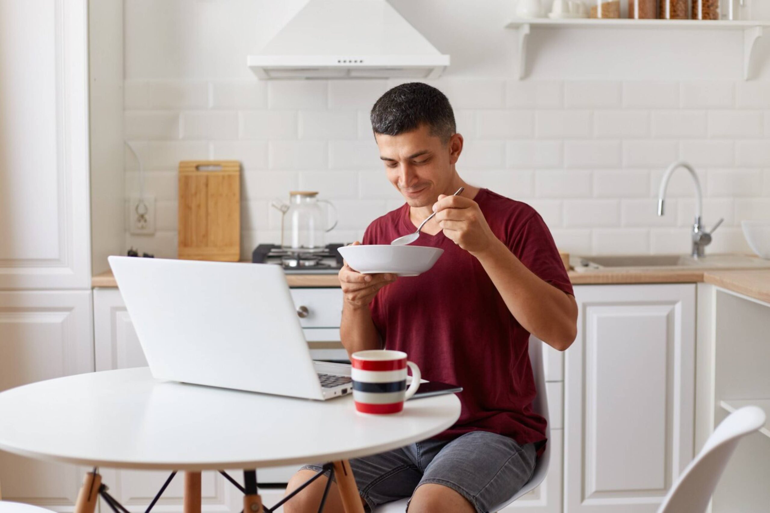 Man eating from a bowl while looking at laptop in a modern kitchen. 