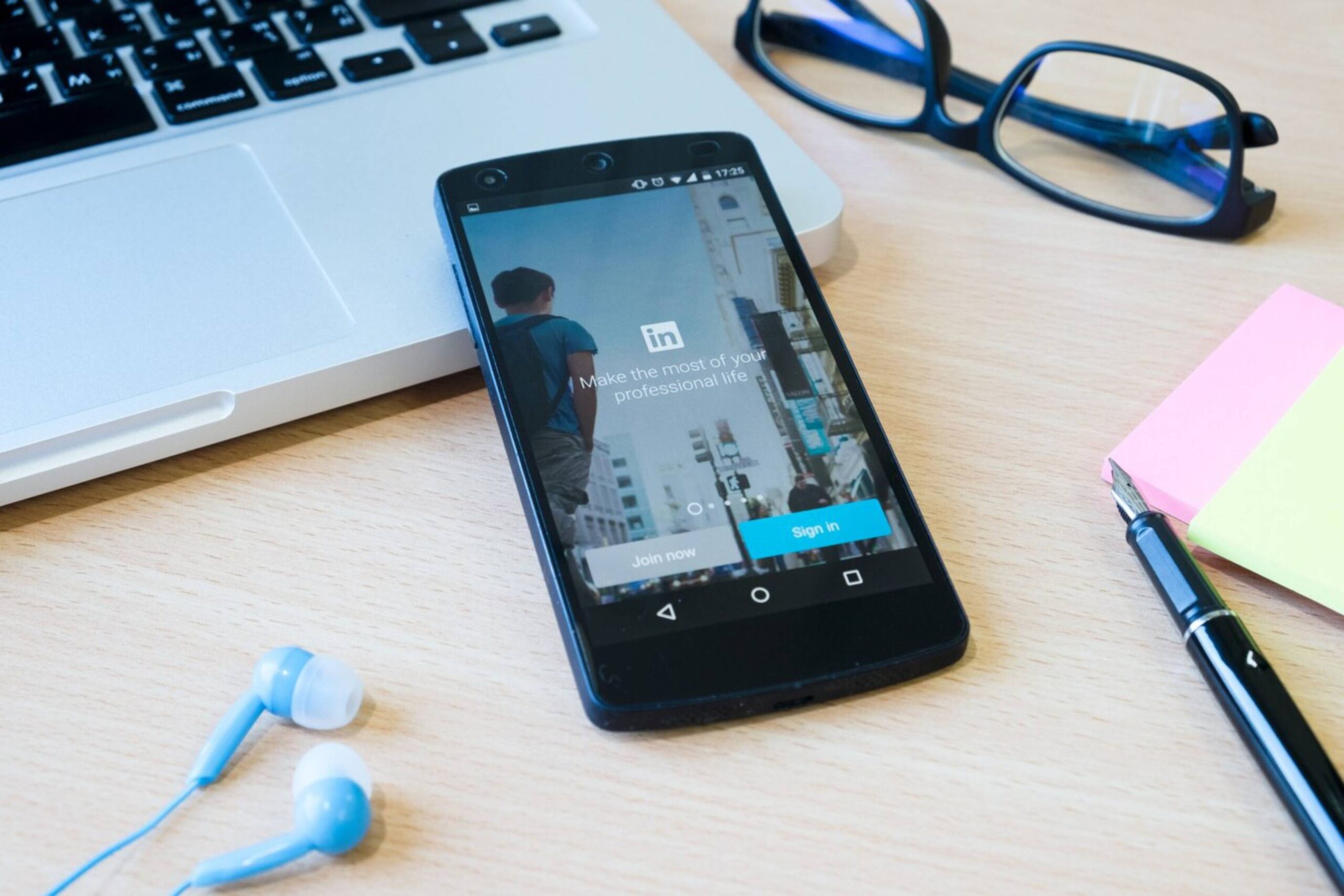 Smartphone displaying LinkedIn login page lies on a desk beside a laptop, glasses, and stationery. 