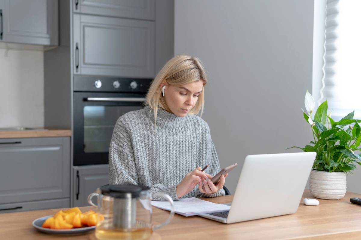 Woman using smartphone at kitchen table with laptop, notepad, and fresh fruit in modern grey kitchen. 