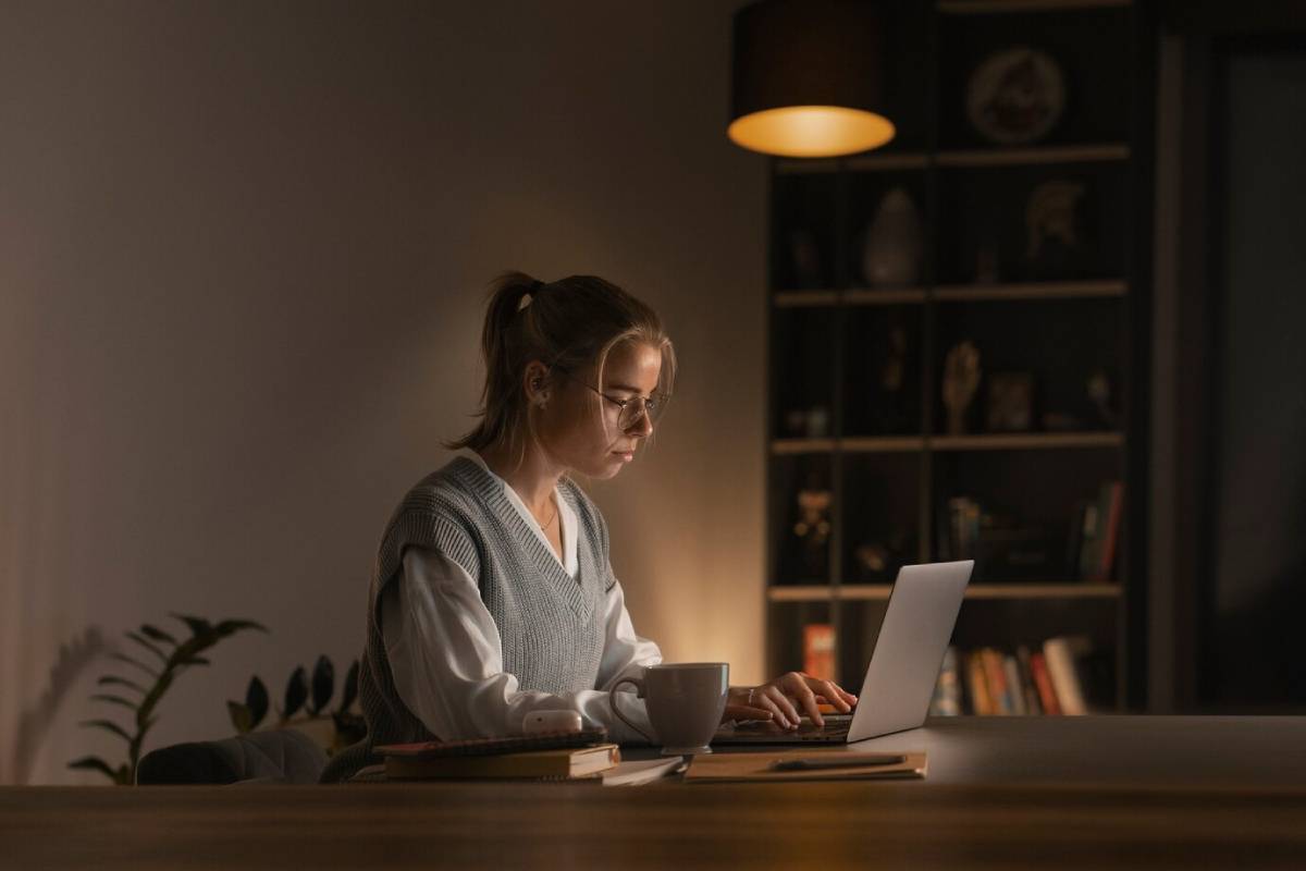  Woman working on laptop at night with soft lighting, surrounded by plants and books.