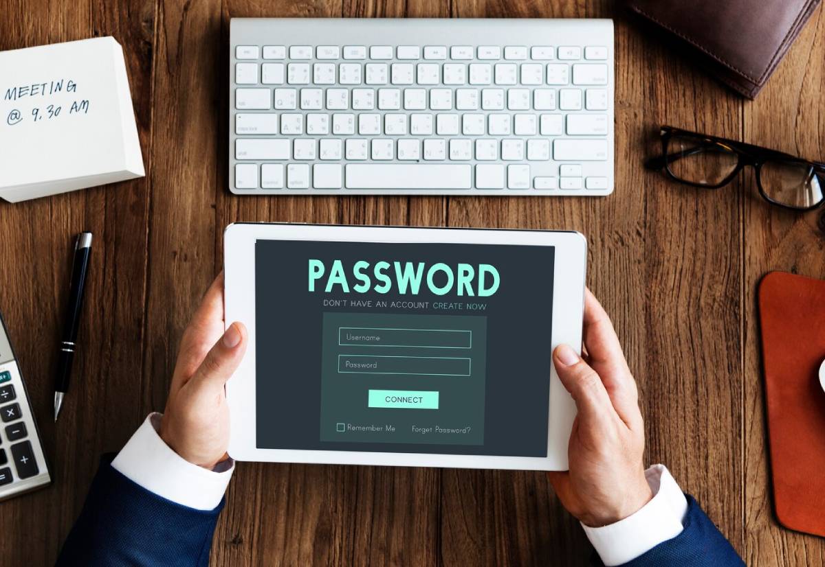 Person holding a tablet displaying a password login screen, with a keyboard and personal items on a wooden desk. 